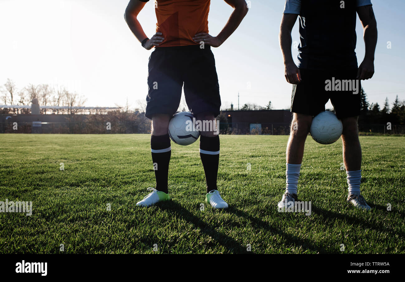 Low section of male friends with soccer balls standing on grassy field against clear sky during sunset Stock Photo