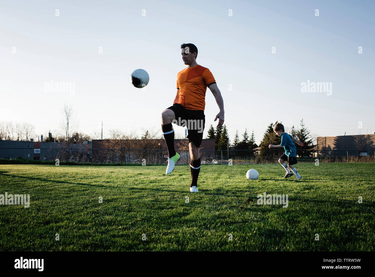Father and son practicing soccer on grassy field against clear sky at park Stock Photo