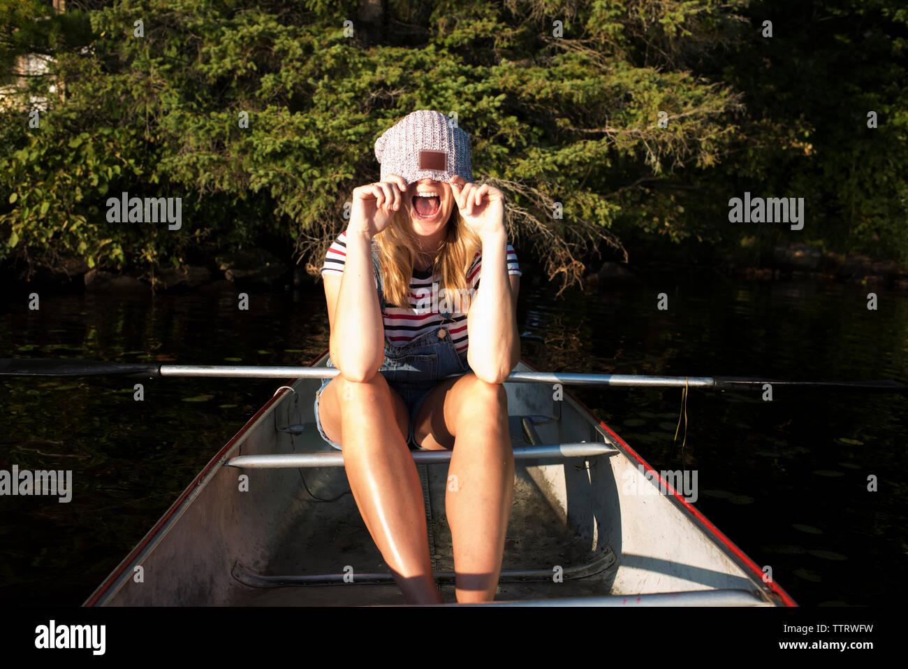 Happy woman covering face with knit hat while sitting in boat on river against trees at Algonquin Provincial Park Stock Photo