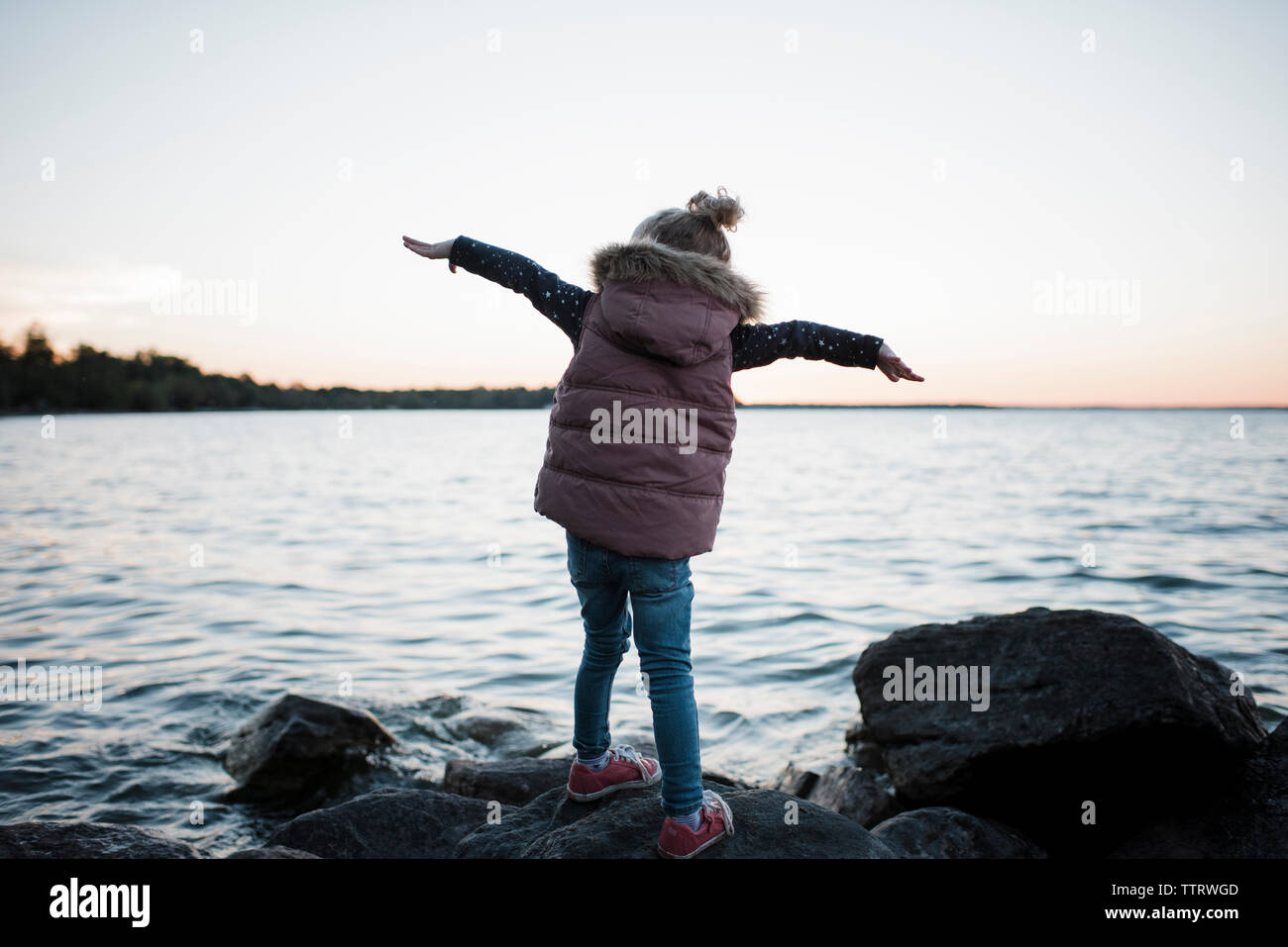 Rear view of girl with arms outstretched standing on rock at lakeshore ...
