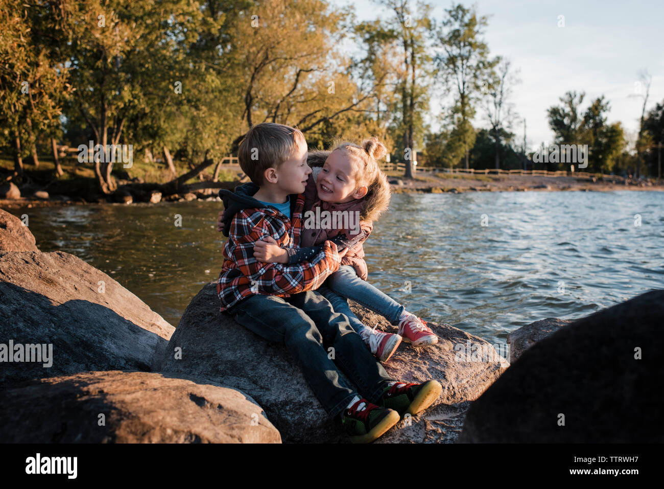 Happy siblings sitting on rock by Lake Simcoe during sunset Stock Photo
