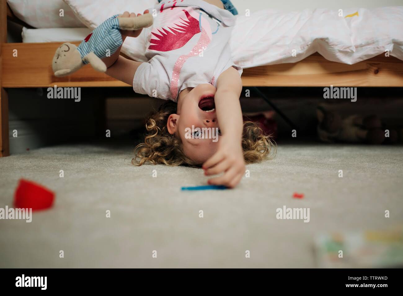 young blonde girl hanging upside down on bed smiling with soft toy Stock Photo