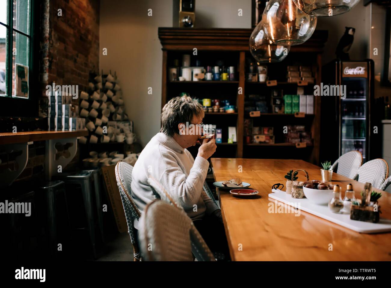 older woman sitting sipping cup of coffee in a cafe and eating bagel Stock Photo