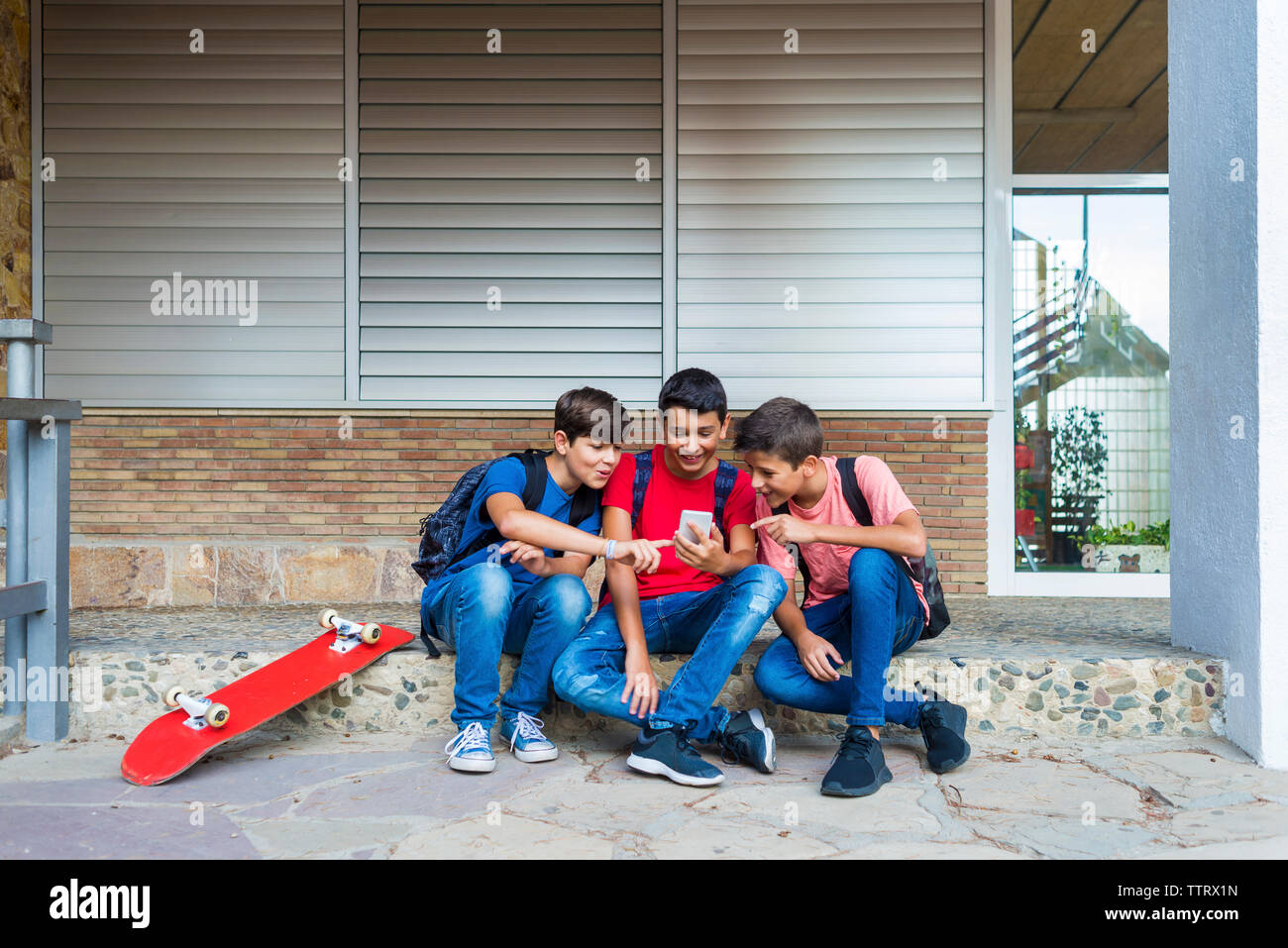 Friends using smart phone while sitting against school building Stock Photo