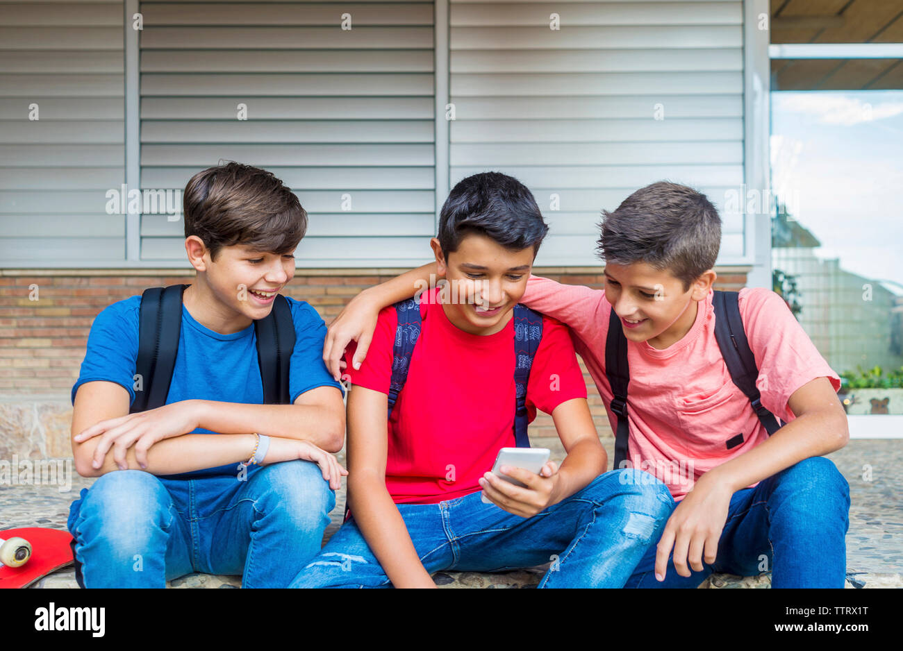 Happy friends using smart phone while sitting against school building Stock Photo