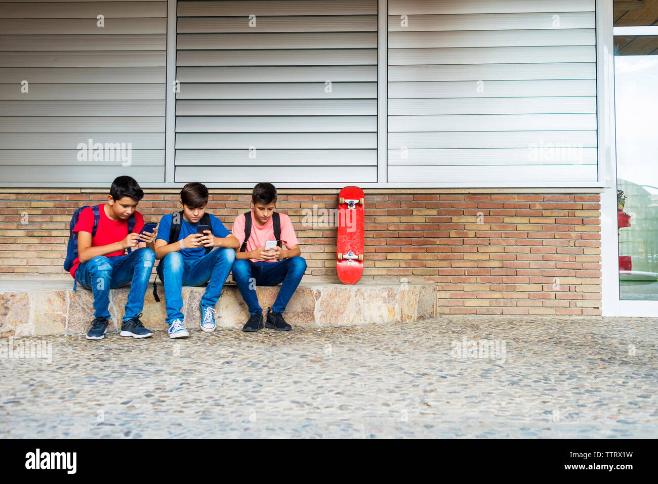 Friends using smart phones while sitting against school building Stock Photo