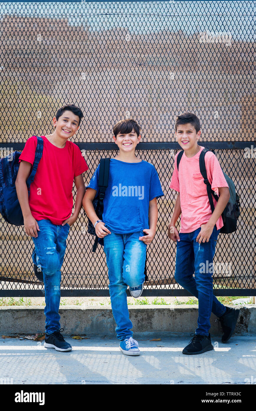 Portrait of smiling friends with backpacks standing by fence Stock Photo
