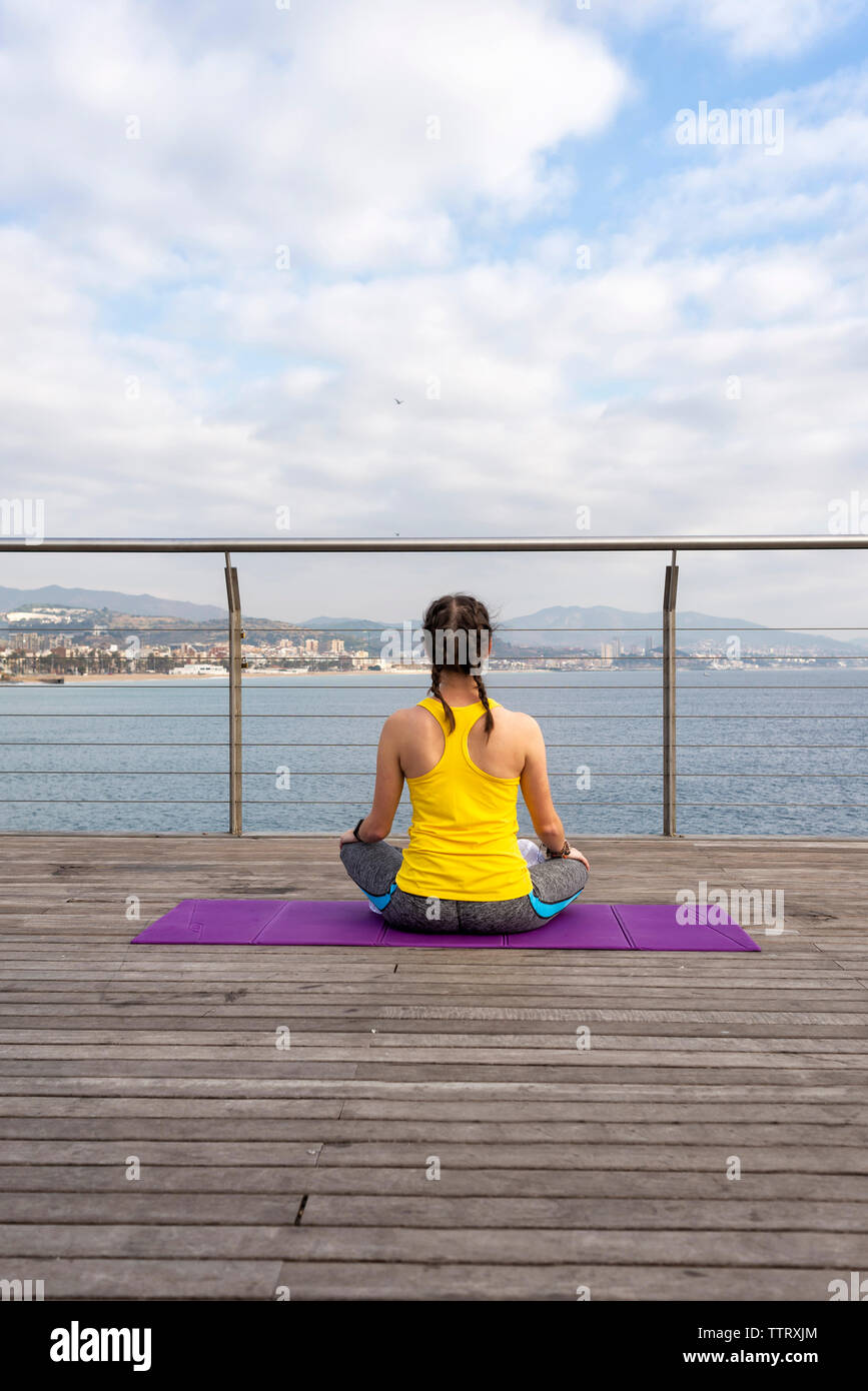 Rear view of woman in sportswear sitting on mat, stretching on pier Stock Photo