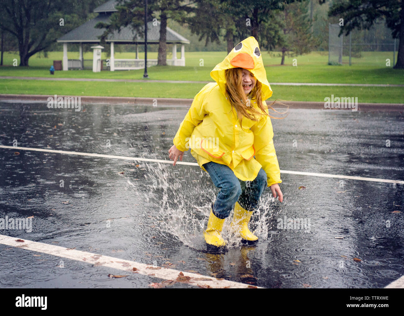 Playful girl wearing raincoat while jumping in puddle during rainfall Stock Photo