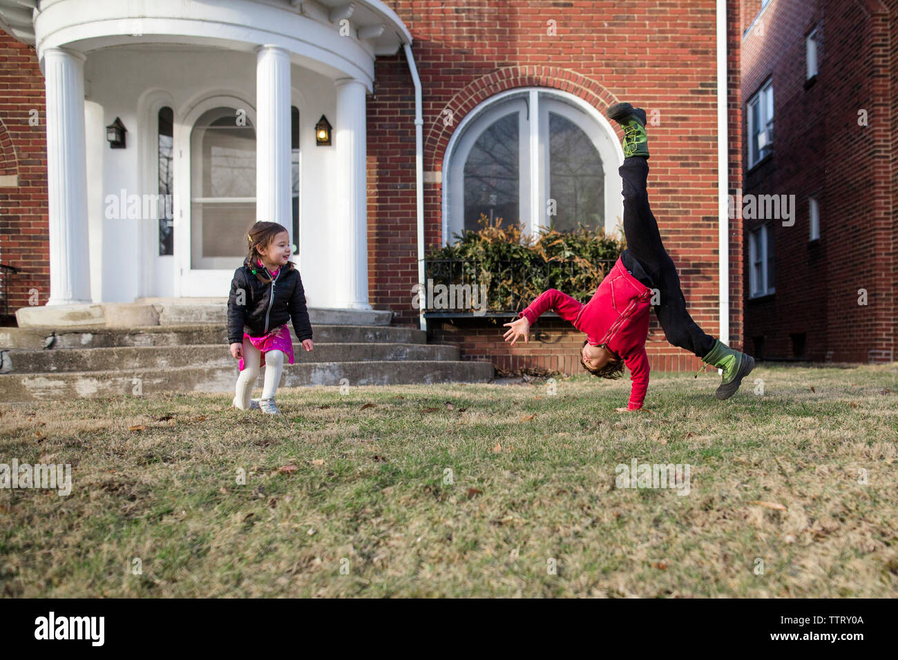 Sister looking at brother practicing cartwheel on lawn against house Stock Photo