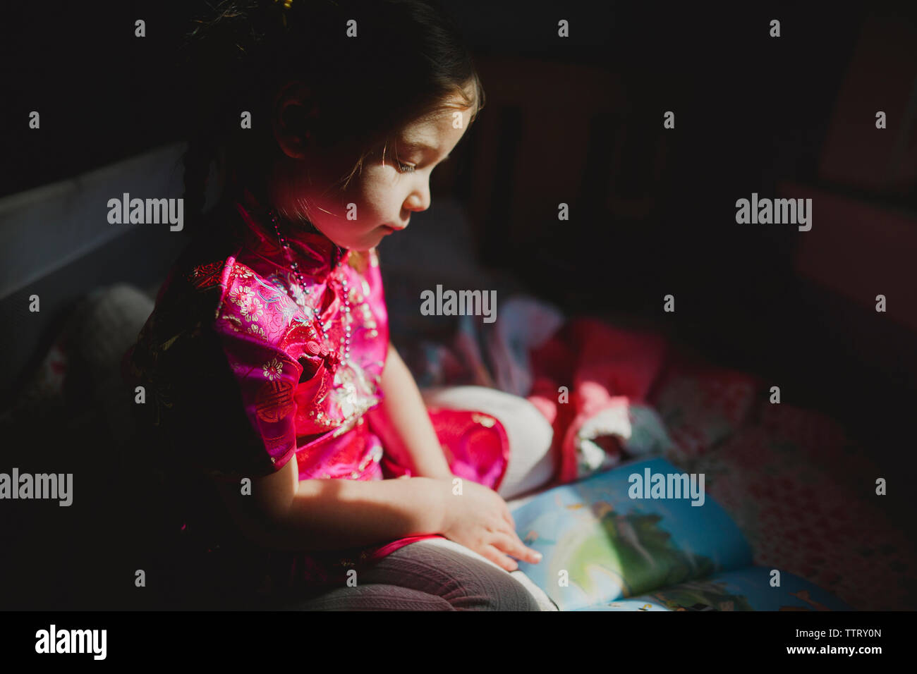 Girl looking at picture book while sitting on bed at home in darkroom ...