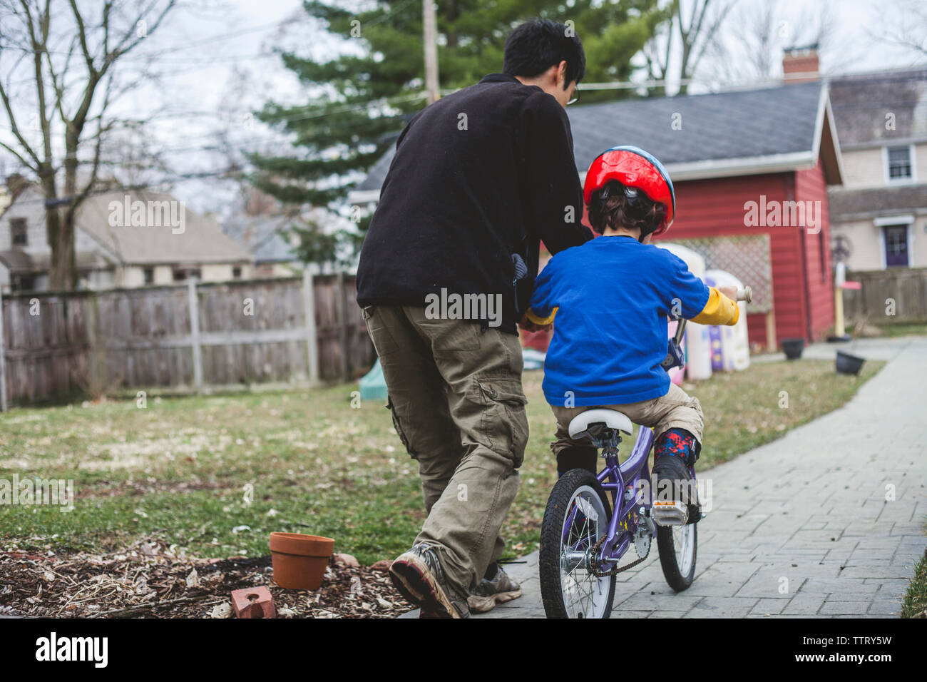 Rear view of father assisting son in riding bicycle on footpath at backyard Stock Photo