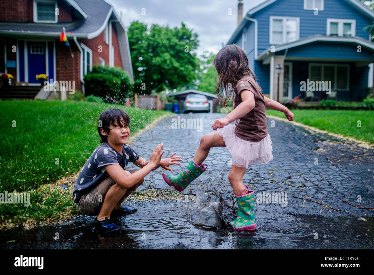 Side view of playful sister splashing puddle on brother during rainy season Stock Photo
