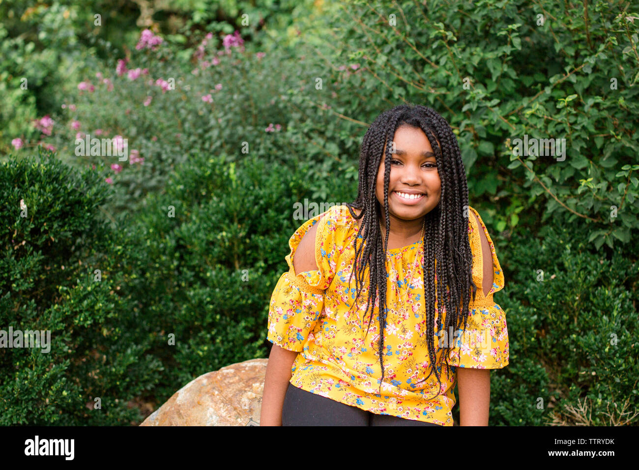 Portrait of smiling girl with braided hair sitting on rock against plants in park Stock Photo