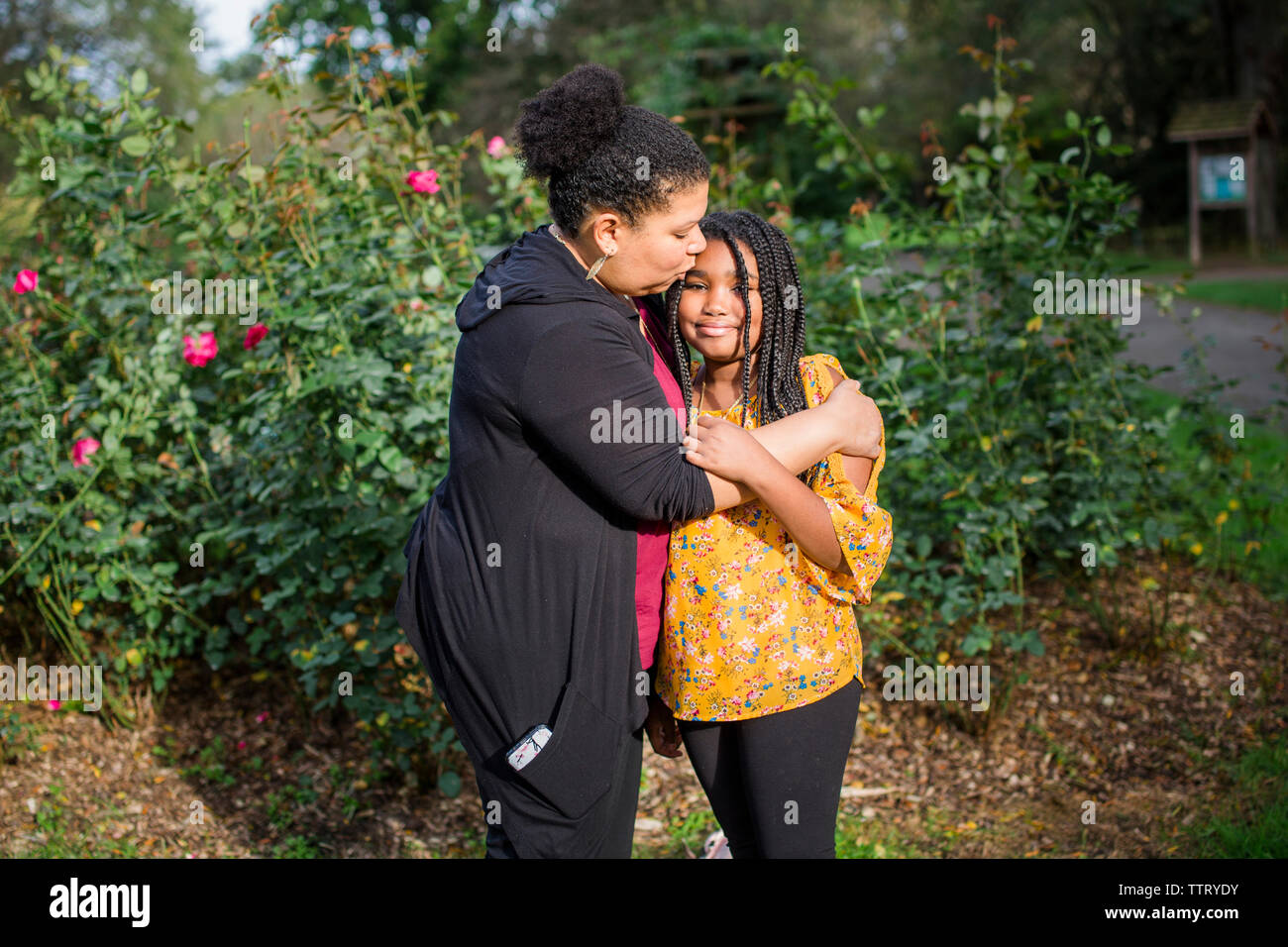 Mother kissing daughter on forehead while standing against plants in park Stock Photo