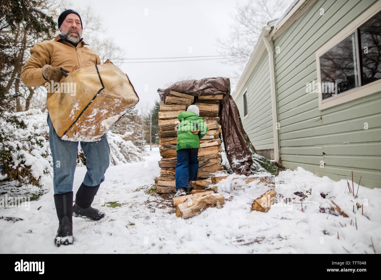 A young child helps his father collect wood from large stack in snow Stock Photo