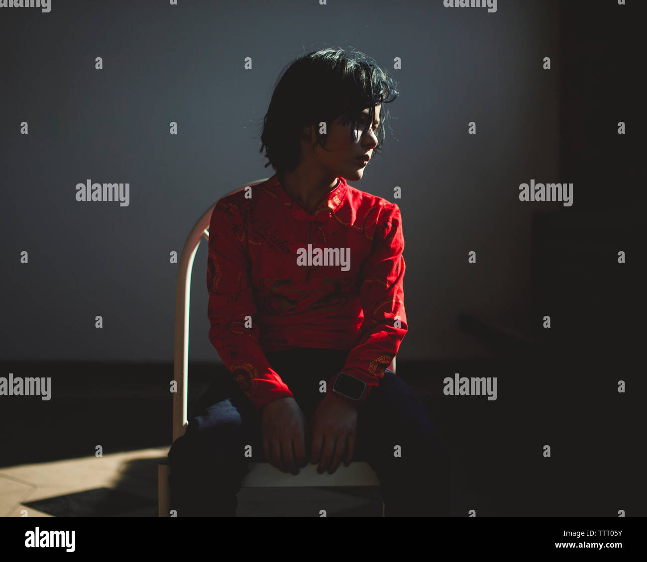 A small beautiful boy sits alone in a dark room bathed in light Stock Photo