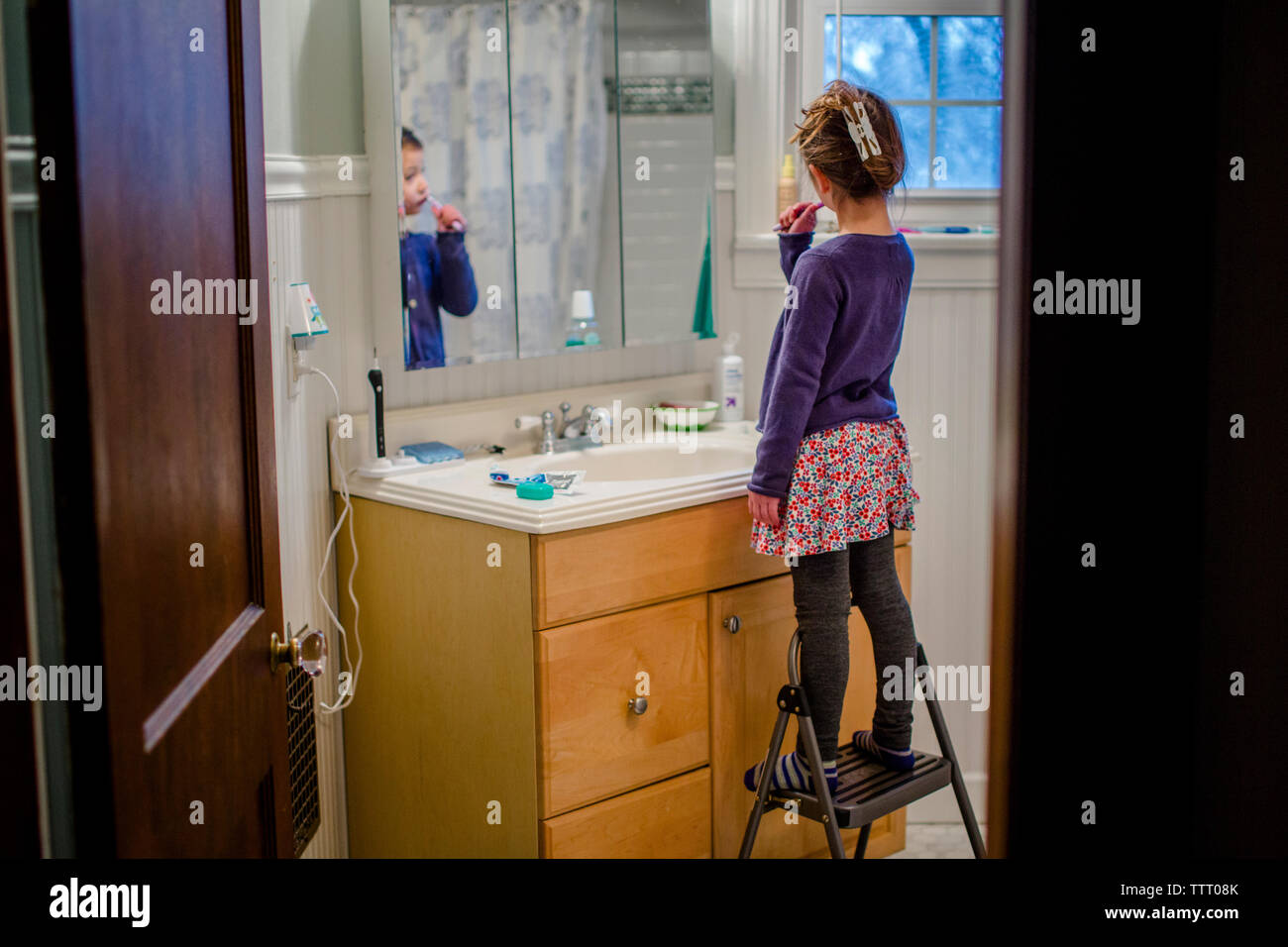 A little girl stands on a stool at the bathroom sink brushing teeth Stock Photo