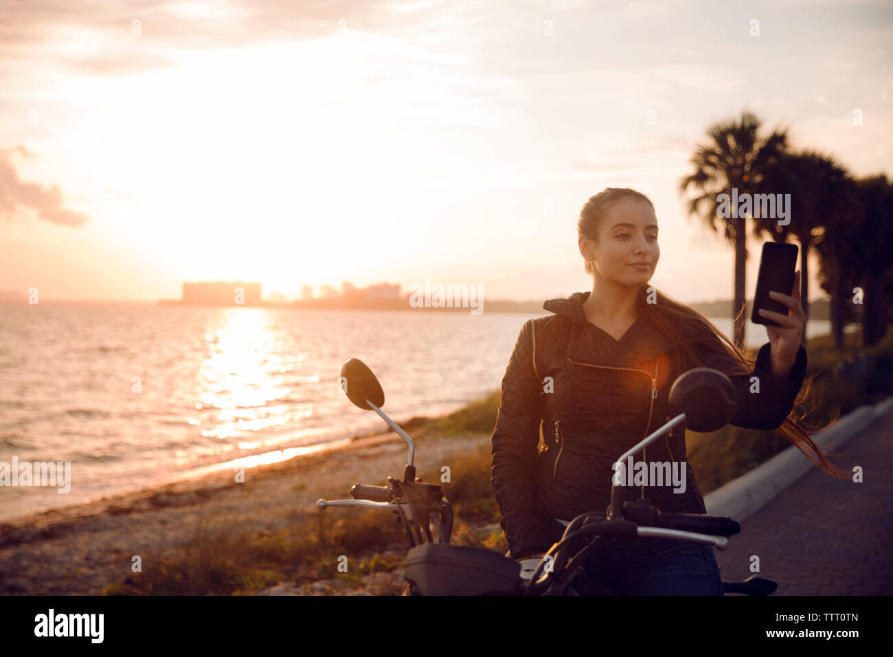 Beautiful woman clicking selfie while sitting on motorcycle during sunset Stock Photo