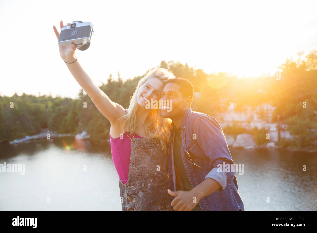 Friends taking selfie through camera by lake during sunset Stock Photo
