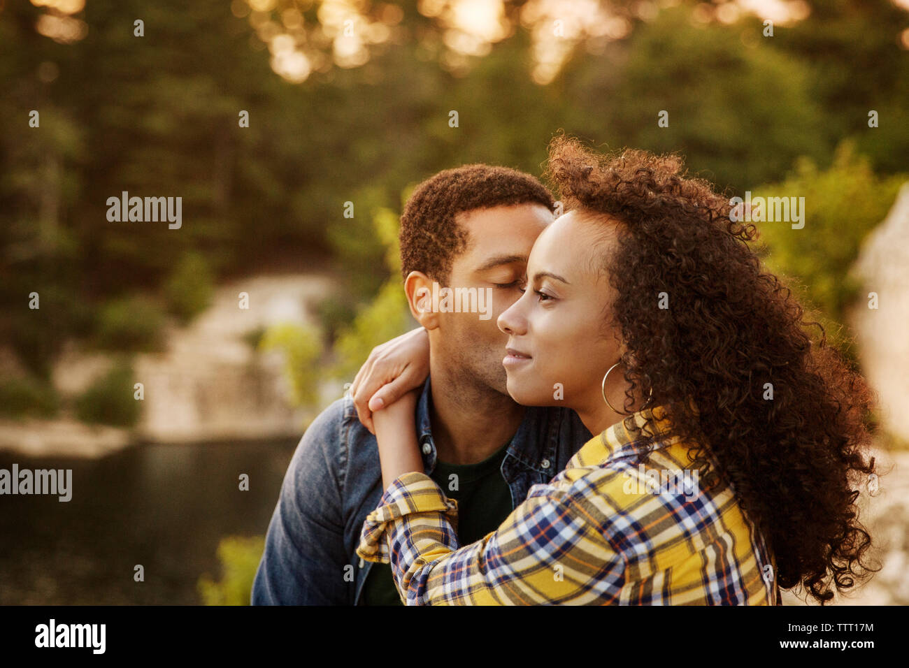 Loving man kissing woman while sitting at lakeshore during sunset Stock Photo
