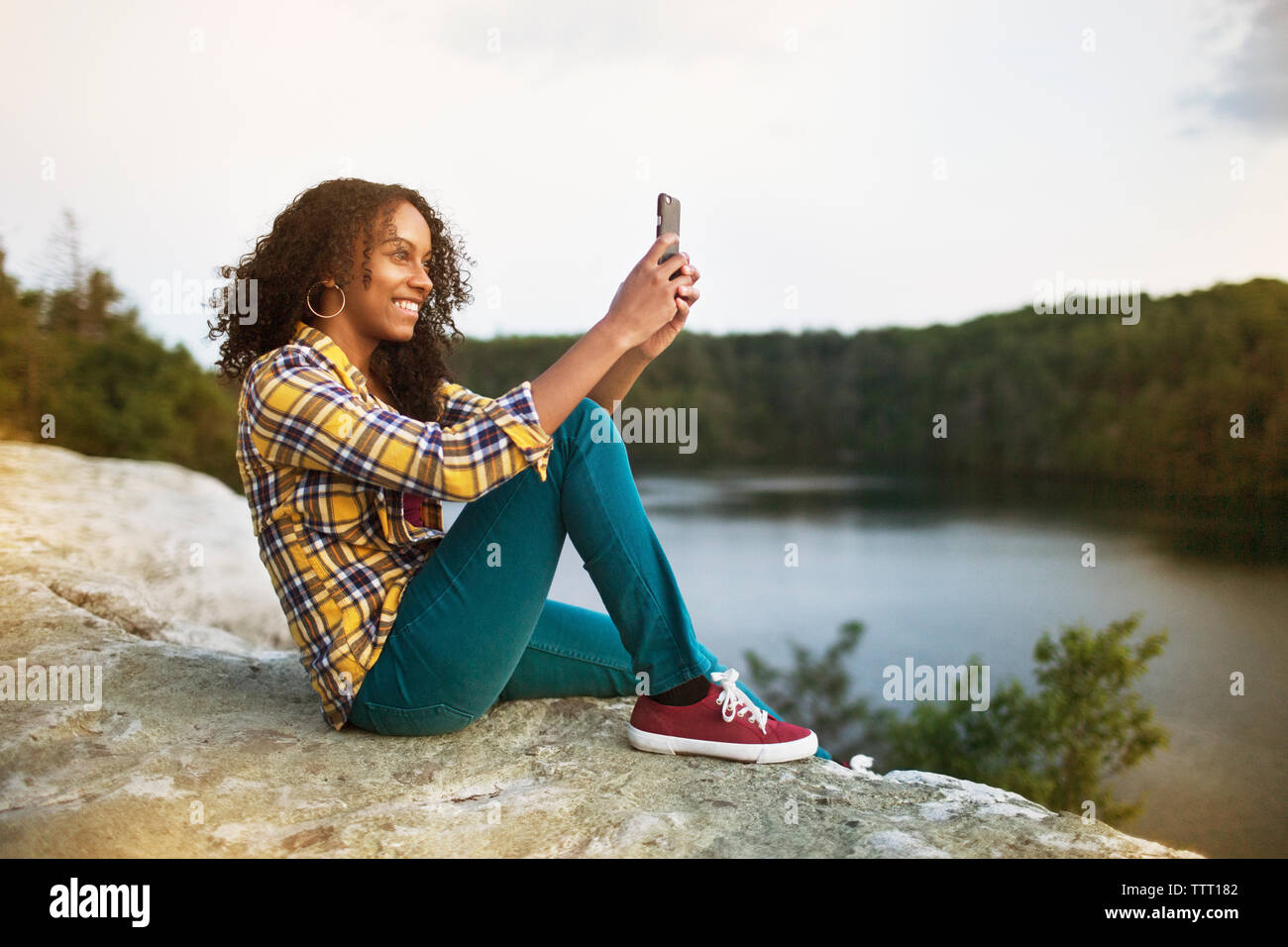 Happy woman taking selfie through smart phone on rock by lake Stock Photo