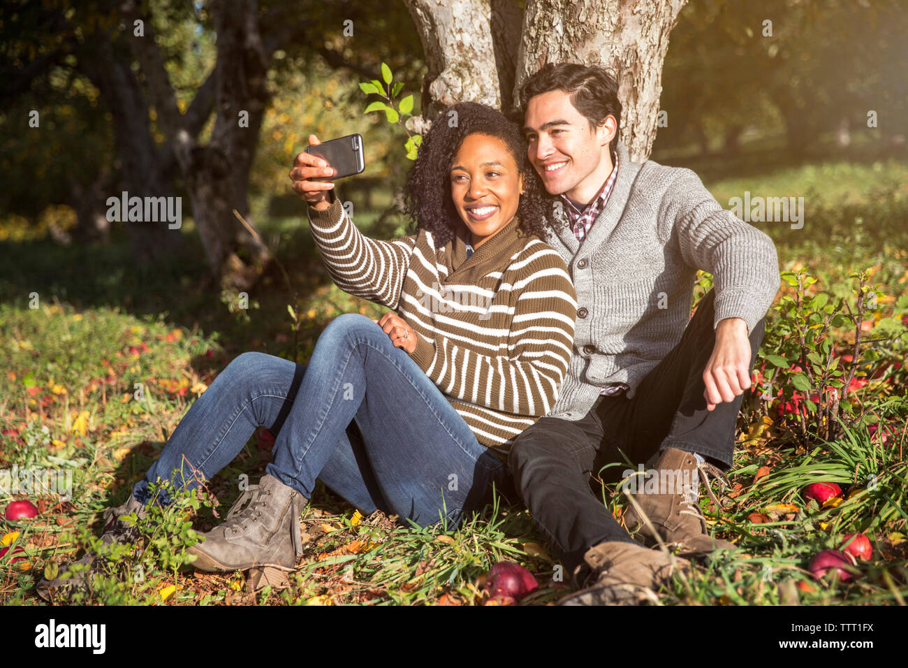 Couple taking selfie while sitting against tree in orchard Stock Photo