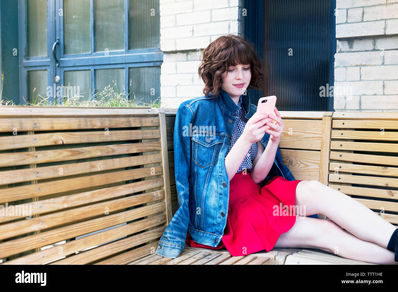 Woman using smart phone while sitting on bench against wall Stock Photo
