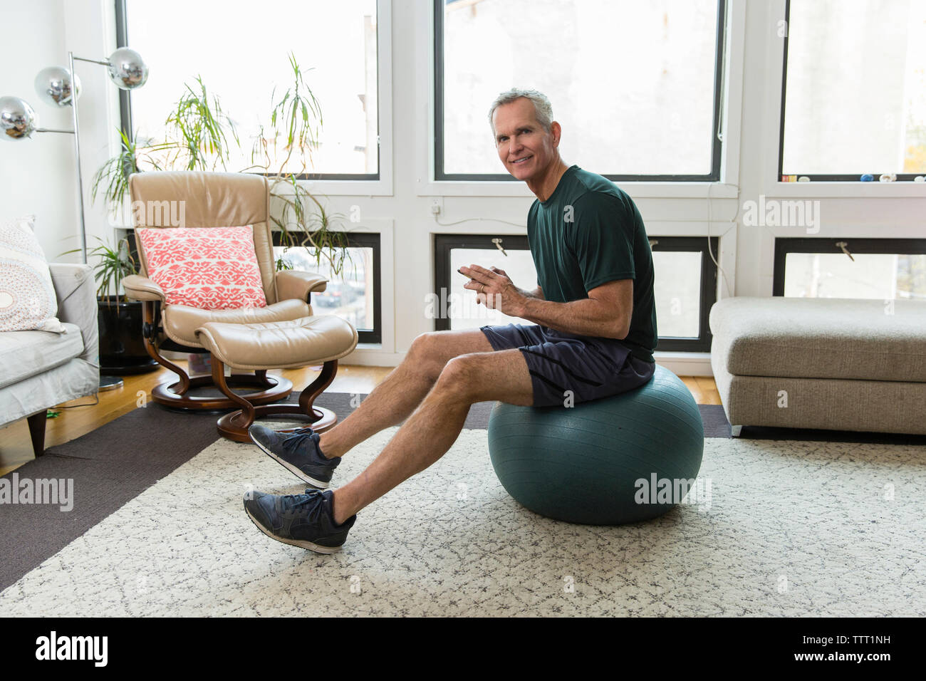 Full length portrait of mature man sitting on fitness ball against windows at home Stock Photo