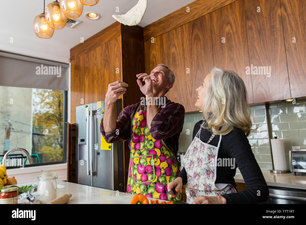 Cheerful man tossing pizza dough while preparing food by woman in kitchen Stock Photo