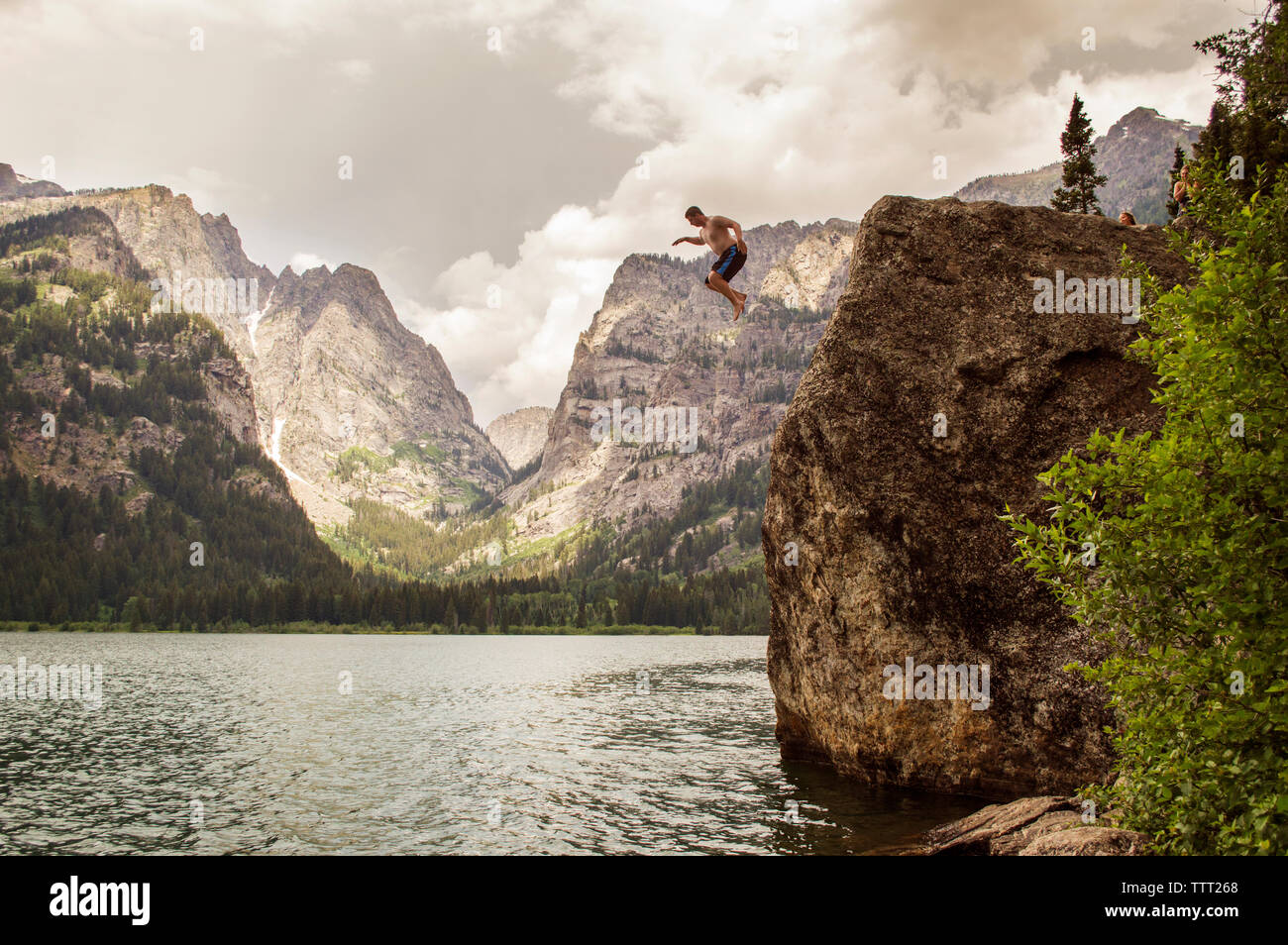 Man jumping from cliff into lake against mountains Stock Photo