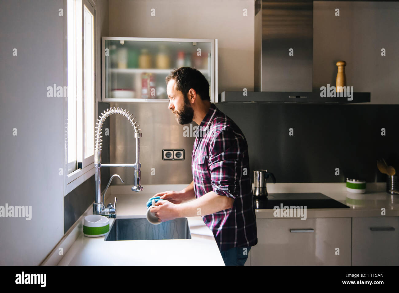 Man with beard and plaid shirt washing dishes at home. Stock Photo