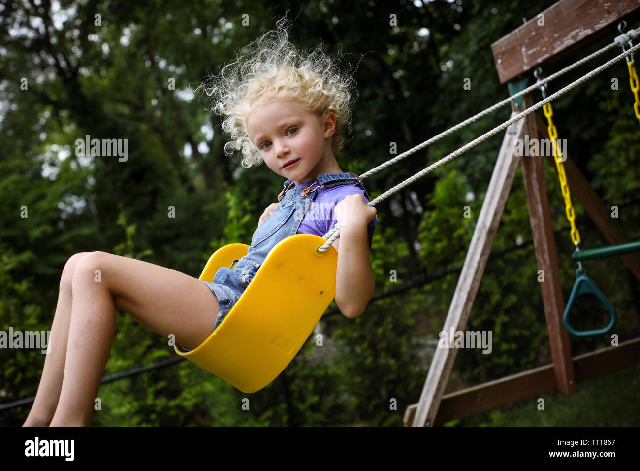 Low angle portrait of cute girl swinging against trees at playground ...