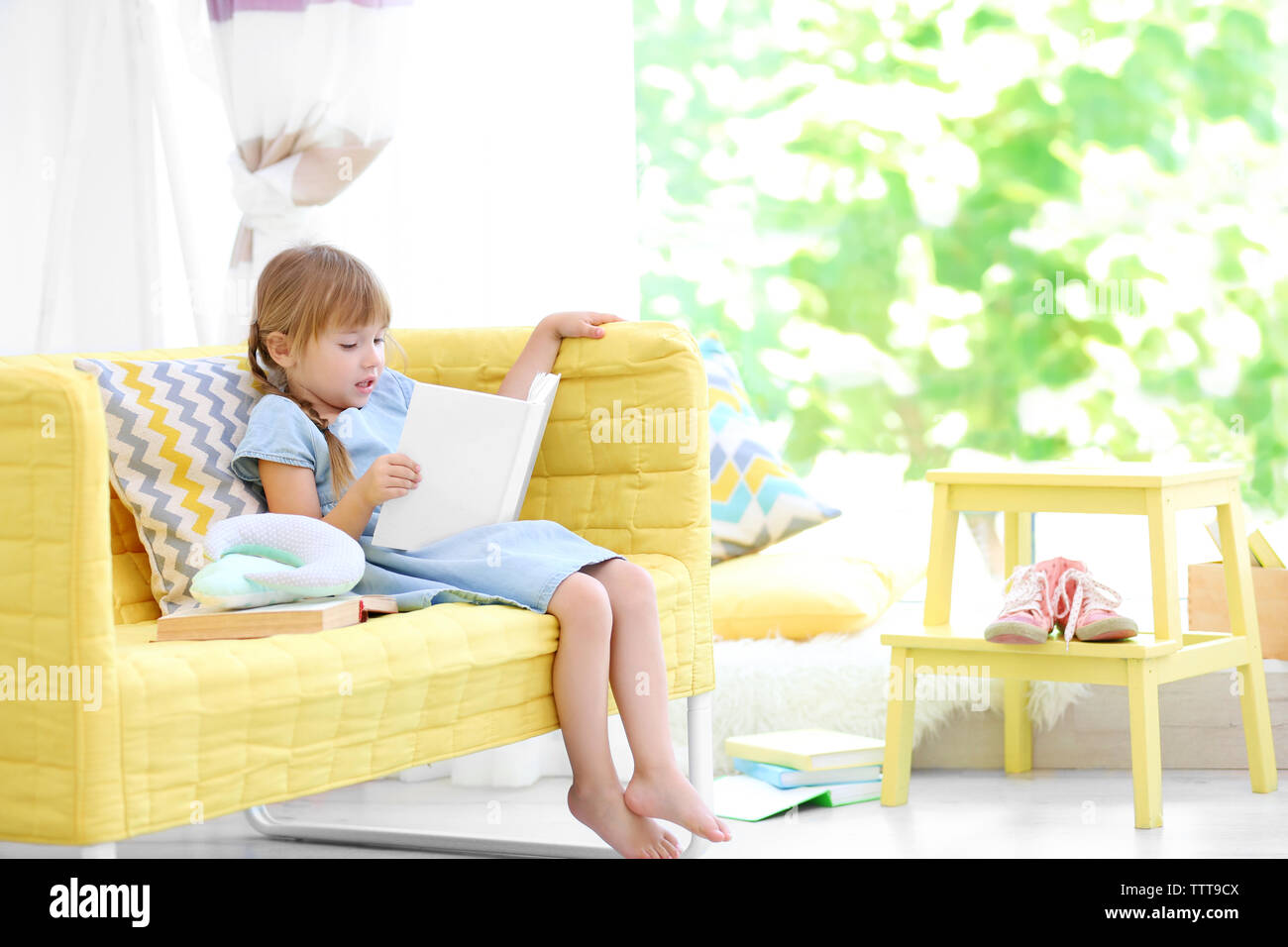 Cute little girl with book sitting on yellow sofa Stock Photo - Alamy