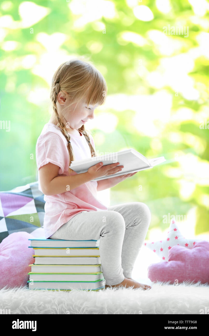 Beautiful little girl sitting on pile of books near window Stock Photo ...