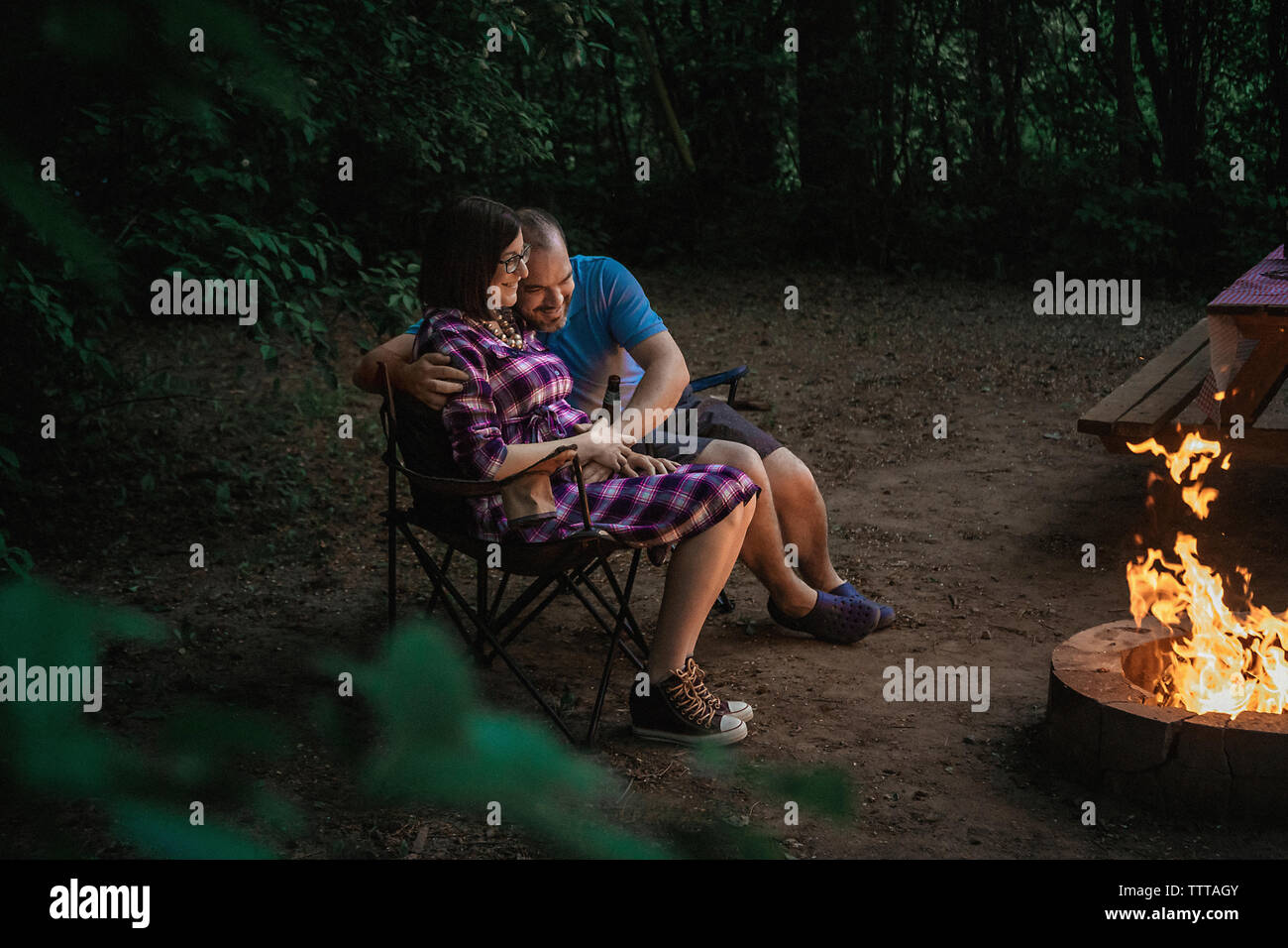 Full length of loving couple sitting on camping chairs by campfire at forest Stock Photo