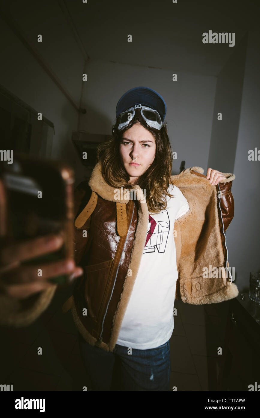 Confident young woman with motorcycle goggles wearing leather jacket while taking selfie in darkroom Stock Photo