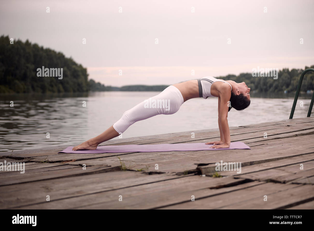 Side view of woman practicing back bridge pose on pier by lake against cloudy sky during sunset Stock Photo