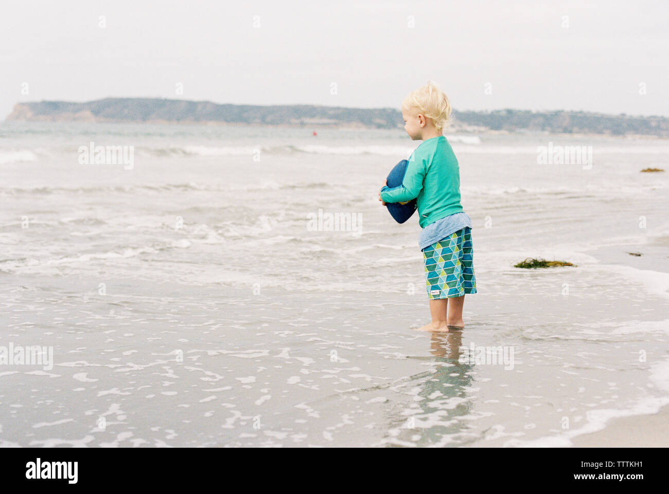 Side view of boy holding American football while standing on shore at beach Stock Photo