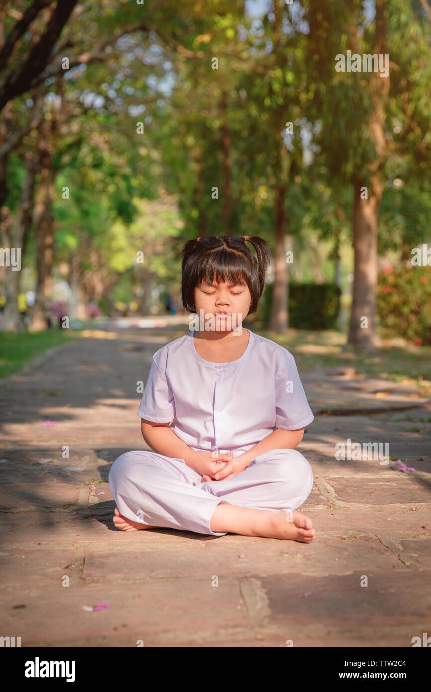 Asian children cute or kid girl sit for meditation with peace and relax in garden pavilion at temple or church and wearing white dress with sunlight o Stock Photo