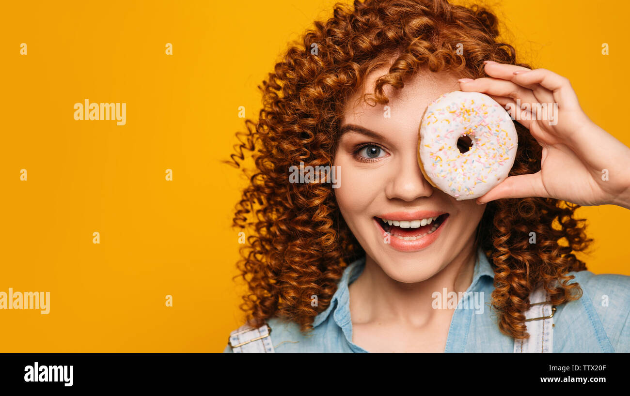 curly red-haired woman holding doughnut near face with surprise emotional face on yellow background Stock Photo