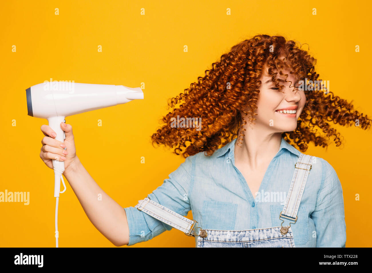 curly red-haired woman using hair dryer on yellow background. Making perfect curls Stock Photo