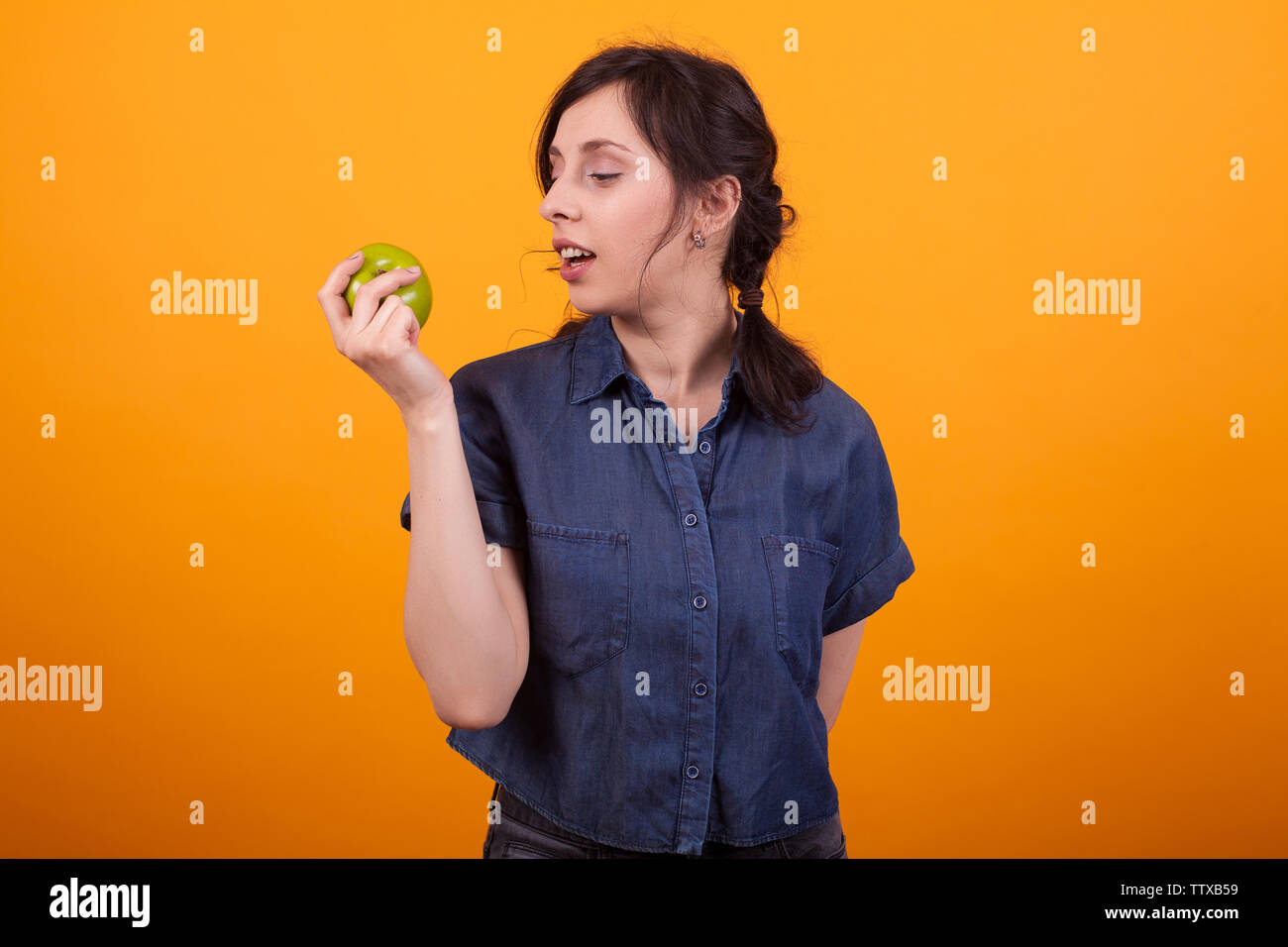Side portrait of cheerful young woman holding and looking at a green apple in studio over yellow background. Woman promoting healthy lifestyle. Eating apples for skin care. Stock Photo
