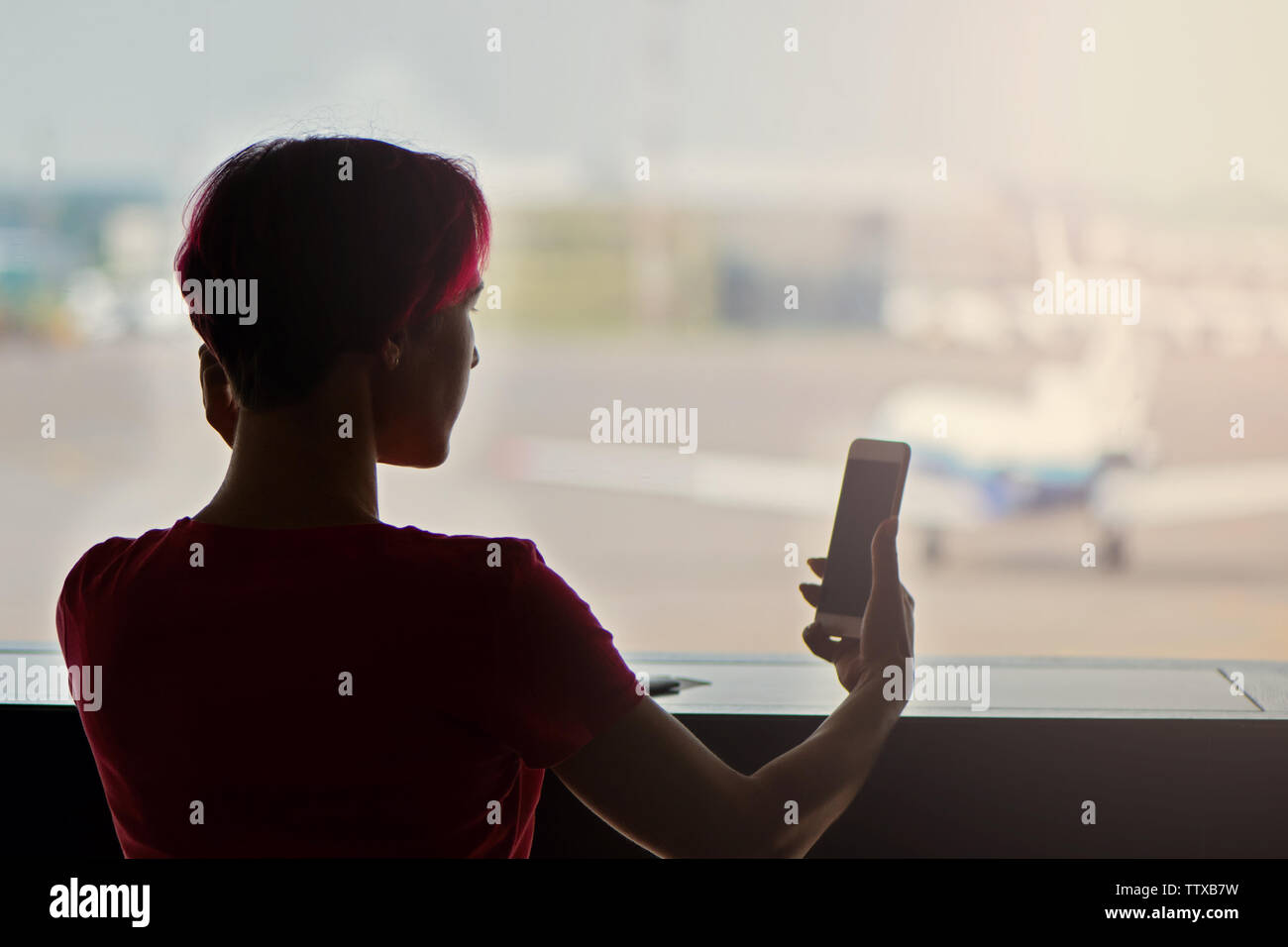 Young woman with pink hair waiting for flying at airport and using smartphone. Back view. Stock Photo