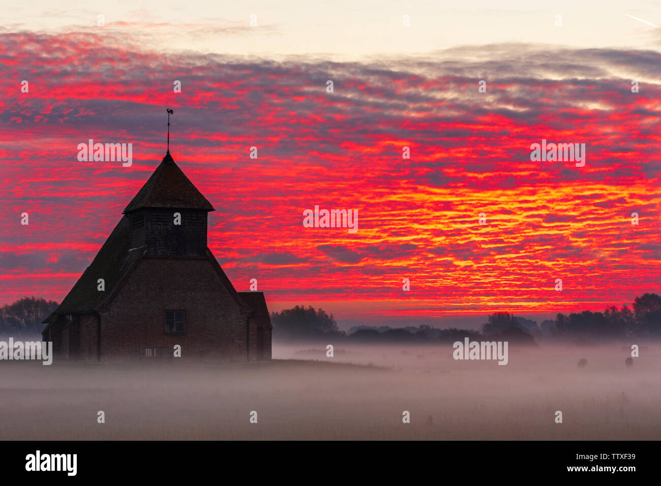 St Thomas Becket church at Romney Marsh. The Church on the marsh at dawn. Ground mist covered, clouds catching red and yellow colours from unrisen sun Stock Photo