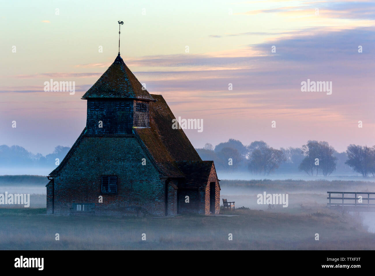 St Thomas Becket church on Romney Marsh. The Church on the marsh at sunup. Sunrise behind clouds, mist on the ground with remote building. Autumn. Stock Photo