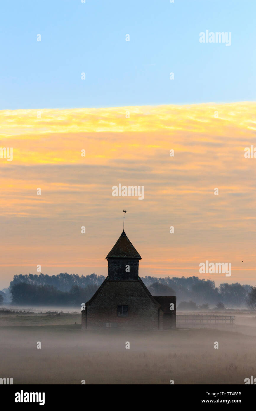 St Thomas Becket church on Romney Marsh. The Church on the marsh at sunup. Sunrise behind clouds, mist on the ground with remote building. Autumn. Stock Photo