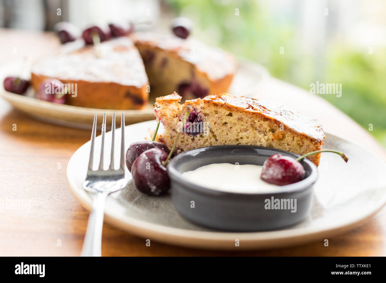 Single slice of cherry cake with icing dusting, cream and cherries on the side, with the rest of the cake in the background Stock Photo