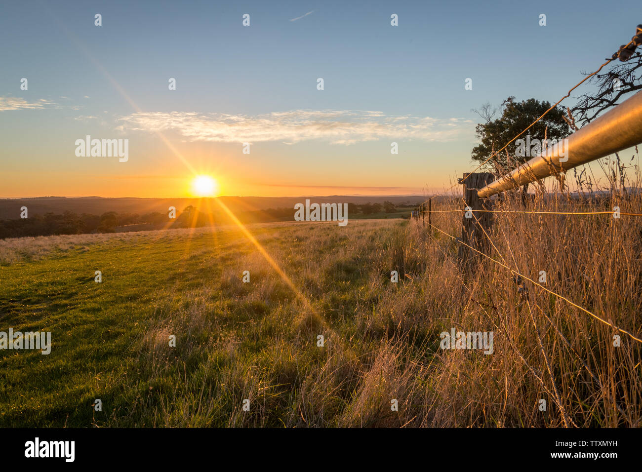 Yellow Sun setting in the distance over hills and paddocks with a farm fence framing the image Stock Photo