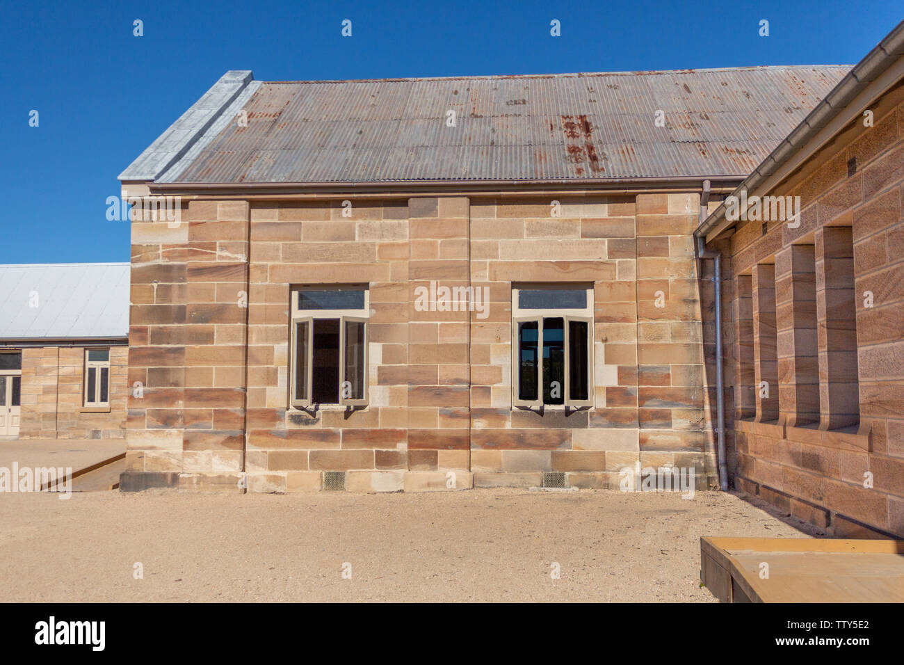 Sandstone convict brick made building with corrugated iron roof, open windows, pebbled courtyard against blue sky Stock Photo